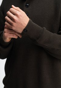 Dunkelbrauner Strickpullover mit gerippter Textur, einem Halsverschluss mit drei Knöpfen und einem markenbezogenen Detail an den Bündchen.