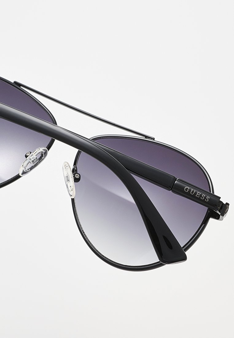 Occhiali da sole Guess con montatura nera, lenti viola sfumate e naselli trasparenti regolabili, mostrati su sfondo bianco.