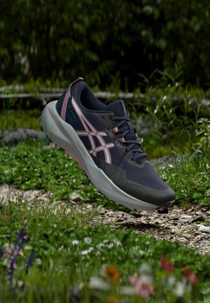 Chaussure de trail running bleu foncé et rose suspendue au-dessus d'un sentier rocheux extérieur avec des plantes vertes et des fleurs sauvages autour.