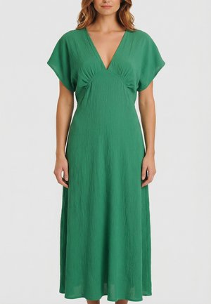 Mujer vistiendo un vestido midi verde con textura, mangas cortas y escote en V profundo, posando contra un fondo blanco liso.