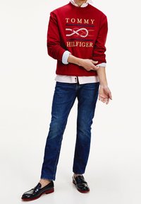 Red sweatshirt with "TOMMY HILFIGER" logo, blue and white striped accents, paired with blue jeans and black loafers with red soles.