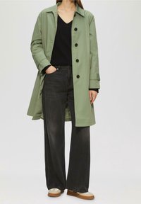 Hellgrüner knie-langer Mantel mit schwarzen Knöpfen, kombiniert mit schwarzen Jeans mit weitem Bein; beige und braune Schuhe. Schwarzes Top mit V-Ausschnitt darunter.