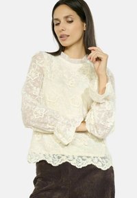 Top bianco in pizzo con motivi floreali e colletto arruffato. Maniche a campana e orlo ondulato. Tessuto testurizzato e leggero.