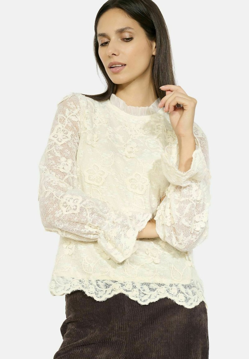 Top bianco in pizzo con motivi floreali e colletto arruffato. Maniche a campana e orlo ondulato. Tessuto testurizzato e leggero.