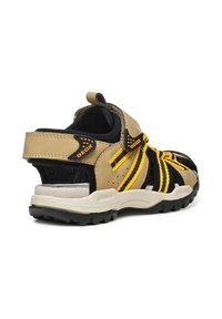 Sandalia en marrón y negro con acentos amarillos, parte superior de malla y cuero, correa ajustable, talón acolchado y suela de goma texturizada para mayor agarre.