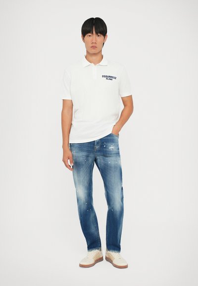 Fehér, rövid ujjú póló sötétkék logóval, kék, koptatott farmernadrággal és bézs sportcipővel, barna gumitalppal párosítva.