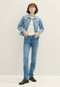 Light blue denim jacket with small front pockets, worn over a white top. Fitted light blue jeans and black shoes complete the outfit.