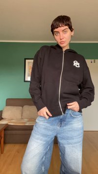 Čierna mikina na zips s prednými vreckami a detailom loga; nosená s svetlomodrými voľnými džínsami. Interiérové prostredie so zelenou stenou a pohovkou.