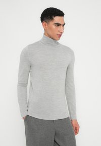 Ung man som bär en ljusgrå polotröja och mörkgrå byxor, stående med händerna i fickorna, ser åt höger mot en vit bakgrund.