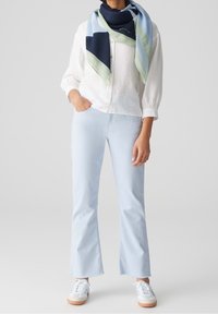 Chemise blanche boutonnée, jean évasé bleu clair, et baskets blanches. Une écharpe multicolore drapée autour du cou, avec des accents bleu marine et vert.