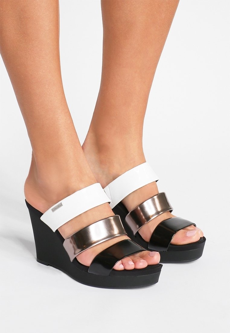 Pieds portant des sandales compensées avec semelles noires et trois brides en blanc, bronze métallique et noir sur les orteils et le cou-de-pied.