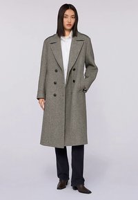 Dvouvrstvý šedý vlněný kabát s širokými klopami, černými knoflíky a bočními kapsami, nošený přes bílou košili a černé kalhoty.