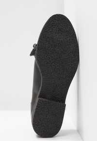 Chaussure en cuir noir reposant semelle contre un mur blanc, montrant la semelle en caoutchouc texturée et le numéro de taille 37 sur le talon.