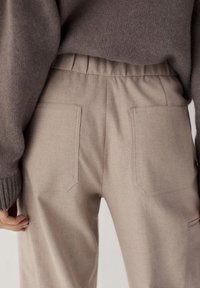 Pantalons beige ajustés avec une finition texturée, dotés de deux poches arrière, d'une taille élastique et d'une coupe décontractée. Portés avec un pull gris.