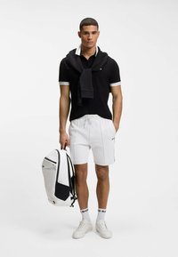 Hombre con polo negro, pantalones cortos blancos y zapatillas, llevando una mochila blanca y negra, con un suéter negro colgado sobre los hombros.