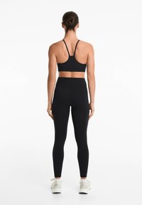 Zwarte sportbeha met dunne bandjes, gecombineerd met hoge zwarte leggings. Model draagt lichtgekleurde sportschoenen. Over het geheel een gladde textuur.