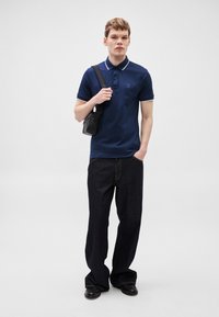 Tricou polo bleumarin cu margini albe, trei nasturi și buzunar aplicat; asortat cu blugi evazați în nuanțe închise și pantofi negri. Geantă mică tip crossbody.