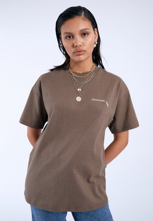 Jonge vrouw met nat uitziend haar draagt een oversized bruine T-shirt en gelaagde gouden en zilveren kettingen tegen een effen achtergrond.