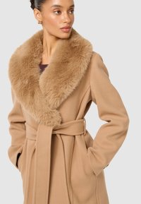 Beige ullrock med bred krage i konstläder, midja med bälte och sidofickor. Mjuk textur med slät finish och detaljerad sömnadsaccent.