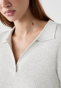 Nahaufnahme des unteren Gesichts und des Halses einer Frau mit Sommersprossen und einem auffälligen Muttermal, die ein hellgraues, geripptes Strickoberteil mit Kragen trägt.