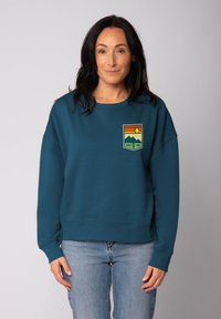 Vrouw met donker haar, gekleed in een teal sweatshirt met een natuurthema "Good Vibe" patch, gecombineerd met lichtblauwe spijkerbroeken, staande voor een neutrale achtergrond.