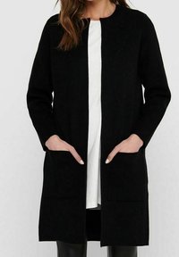 Dlouhý černý kardigan z měkké látky, s rovnými liniemi, otevřeným předním dílem a dvěma předními kapsami. Nositý přes bílý top.