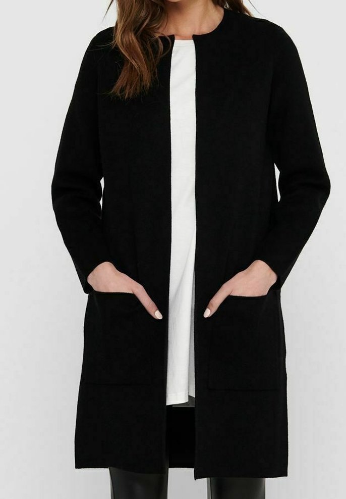 Dlouhý černý kardigan z měkké látky, s rovnými liniemi, otevřeným předním dílem a dvěma předními kapsami. Nositý přes bílý top.