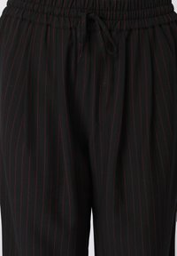 Sorte pinstripe bukser med en tekstureret finish, der har en elastisk talje med rynket effekt og snøre, detaljeret med kontrasterende røde striber.