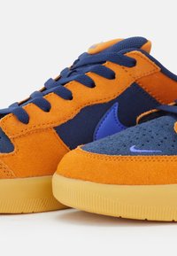 Sneakers med orange mocka och marinblått tyg på ovandelen, med lila detaljer. Texturvariation mellan materialen; gummisula med räfflor.