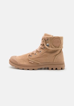 Bottes chelsea en toile beige avec un embout en caoutchouc, des œillets en métal et un patch logo sur le col. Surface texturée et semelle robuste.