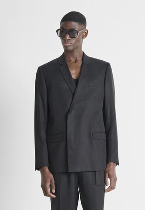 Man met een oversized zwarte blazer en bijpassende broek, geaccentueerd met grote zwarte zonnebrillen, staand tegen een effen witte achtergrond.
