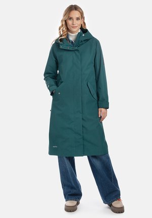 Vrouw draagt een lange teal kleurige hooded jas over een witte coltrui en wijde blauwe jeans, staand met één hand in de zak.