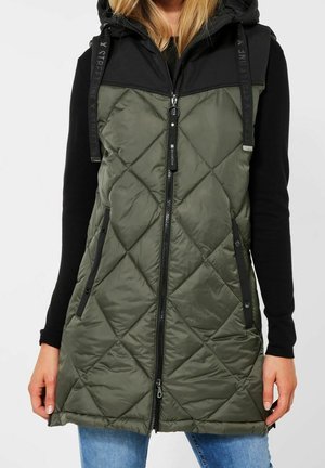 Kvinde iført en ærmeløs, quiltet olivengrøn vest med sort hætte, lynlås og sidelommer over en sort langærmet top og blå jeans.