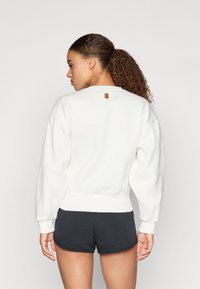 Λευκή μπλούζα με ραμμένο λαιμό και μανσέτες, με ένα μικρό πολύχρωμο λογότυπο στο πίσω μέρος. Συνδυάζεται με σκούρα σορτς.