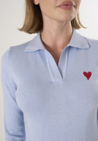 Polo shirt lavorato a maglia di colore azzurro chiaro con colletto e un'applicazione a forma di cuore rosso sul lato sinistro del petto. Tessuto morbido e design aderente.