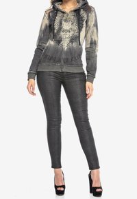 Grijze hoodie met geborduurd leeuwmotief, gecombineerd met glanzende zwarte skinny jeans. De hoodie heeft een capuchon met trekkoord en verweerde randen.