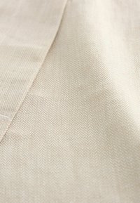 Beige herringbone-kangas, jossa on hienovarainen tekstuuri, diagonaalinen kuvio ja sileä pinta. Vaalea väri parantaa kuvion näkyvyyttä.