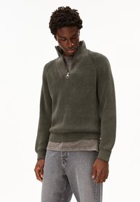 Ein Mann trägt einen olivgrünen, gestreiften Strickpullover mit einem halben Reißverschlusskragen, der über einem verblassten grauen T-Shirt getragen wird, kombiniert mit hellgrauen Jeans.