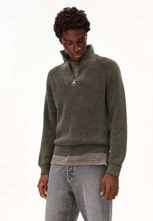 Homme portant un pull en maille côtelée vert olive avec un col zippé, superposé à un t-shirt gris délavé, assorti à un jean gris clair.