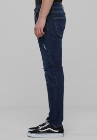 Jeans de mezclilla azul oscuro con un ajuste slim, con detalles desgastados y costuras visibles. Combinados con zapatillas negras de caña baja.