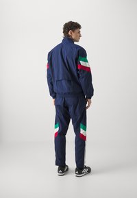 Námornícky teplákový komplet s farebným blokovaním v červenej, zelenej a bielej farbe. Ľahký materiál, elastické manžety a voľný strih. Čierne športové topánky.