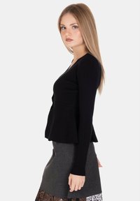 Giovane donna con lunghi capelli biondi, che indossa un top nero a maniche lunghe e una gonna grigia con bordi in pizzo nero, in posizione laterale.