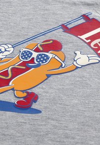 Κινούμενο σχέδιο με λουκάνικο που έχει χέρια, πόδια, κόκκινα παπούτσια και γυαλιά ηλίου με σχέδιο αστεριών, κρατώντας μια κόκκινη σημαία με λευκό κείμενο σε γκρι υφασμάτινο φόντο.