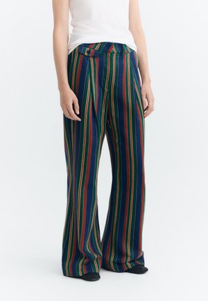 Personne portant un pantalon à jambes larges avec des rayures verticales vertes, bleues, rouges et jaunes, associé à un haut blanc et des chaussures noires.