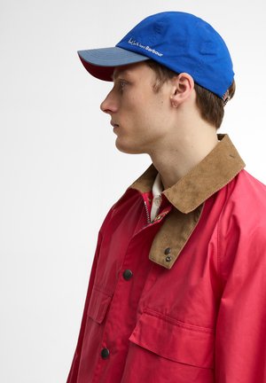 Νεαρός άνδρας με μπλε και γκρι καπέλο, κόκκινο μπουφάν με καφέ γιακά από κορδόνι, και λευκό πουκάμισο, εμφανίζεται σε προφίλ με φόντο ανοιχτόχρωμο.