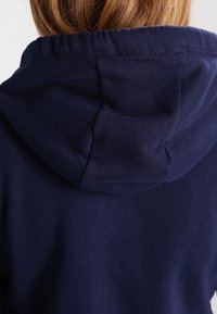 Marineblå hoodie med blød tekstur, der har en snøre i hætten og et enkelt, rent design. Stoffet ser let og behageligt ud.