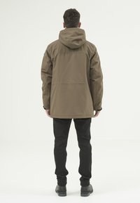 Brun vattentät jacka med huva, med en rak skärning och elastiska ärmar, parat med svarta byxor och stövlar.