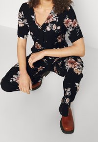 Osoba klęczy, ubrana w czarną kwiatową bluzkę i spodnie, zestawione z brązowymi butami na kwadratowym obcasie, na tle jednolitej ściany.