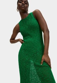 Grünes gehäkeltes Kleid mit ärmellosem Design, das ein strukturiertes Muster und einen dezenten Seitenschlitz aufweist. Der Stoff wirkt leicht und atmungsaktiv.