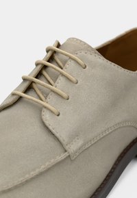 Μπεζ σουέτ παπούτσι με κορδόνια και λεία υφή, διαθέτοντας έξι τρύπες για κορδόνια, ραμμένη τελειώματα και καφέ καουτσούκ σόλα.